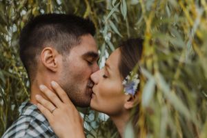 Couple sharing a romantic kiss surrounded by lush green leaves outdoors.
