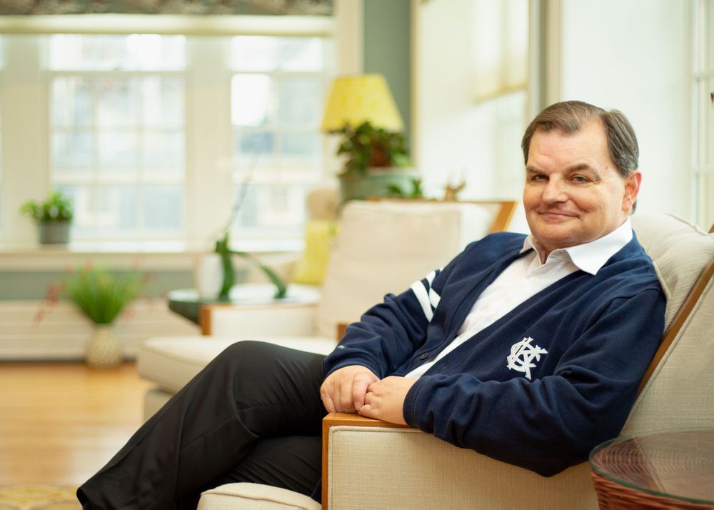 A man wearing a dark navy blue suit lounges on a white chair.