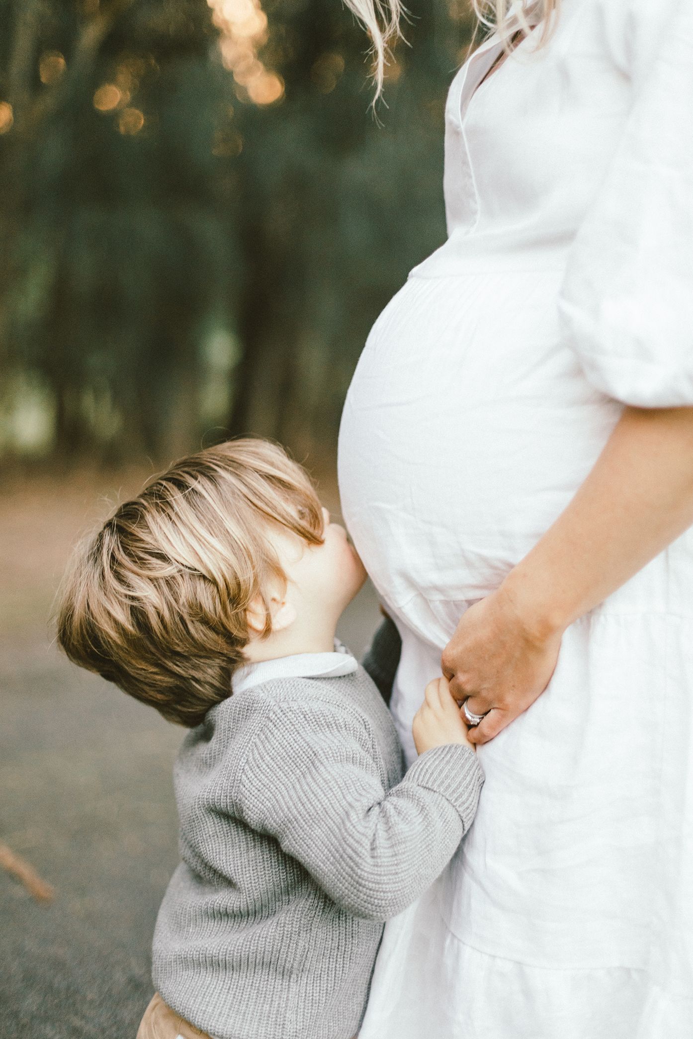 Young child in gray sweater hugs an expectant mother wearing a white dress in scenic outdoor setting.