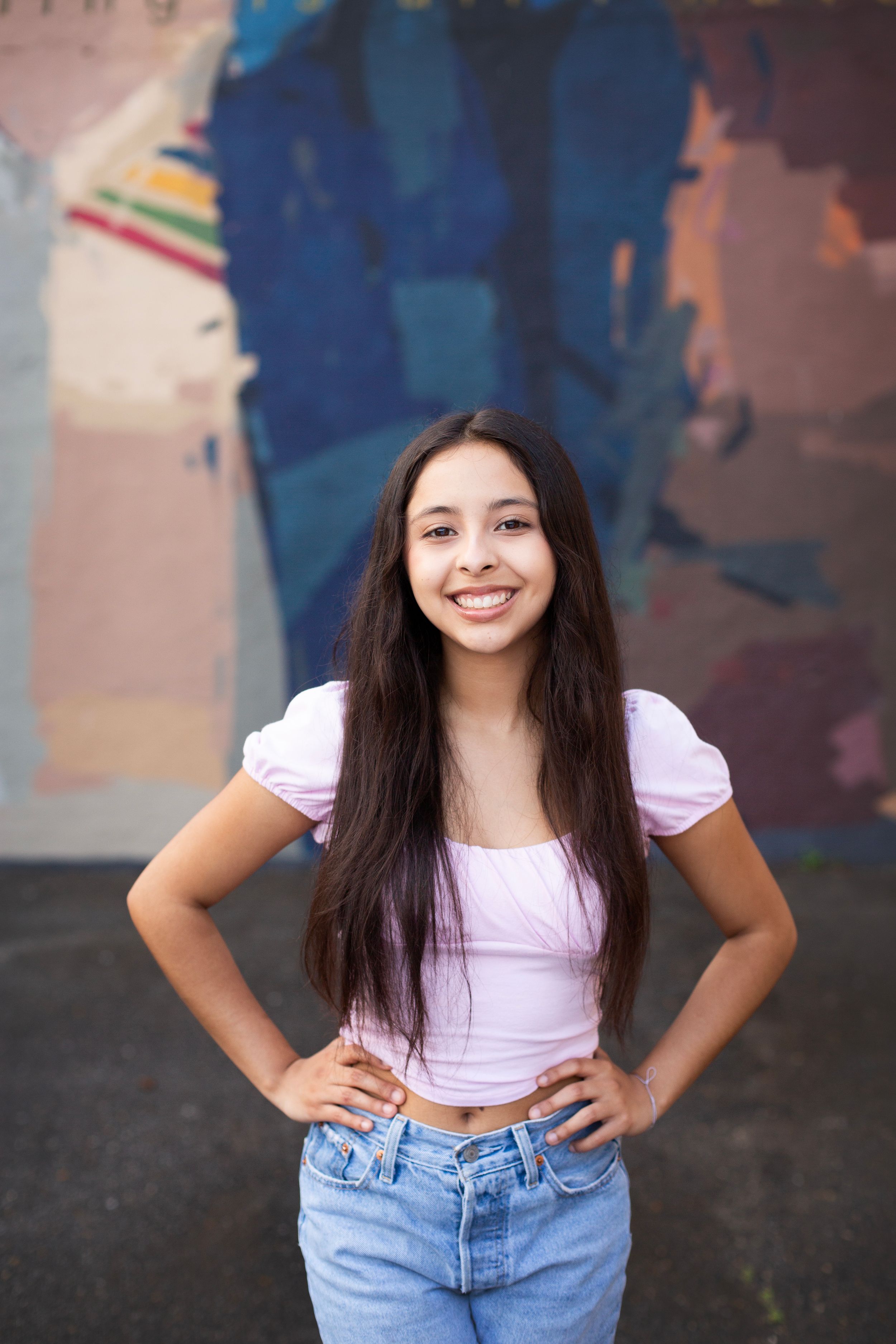 High School Senior girl wearing a pink shirt and jeans looking at the camera with both hands on her hips.