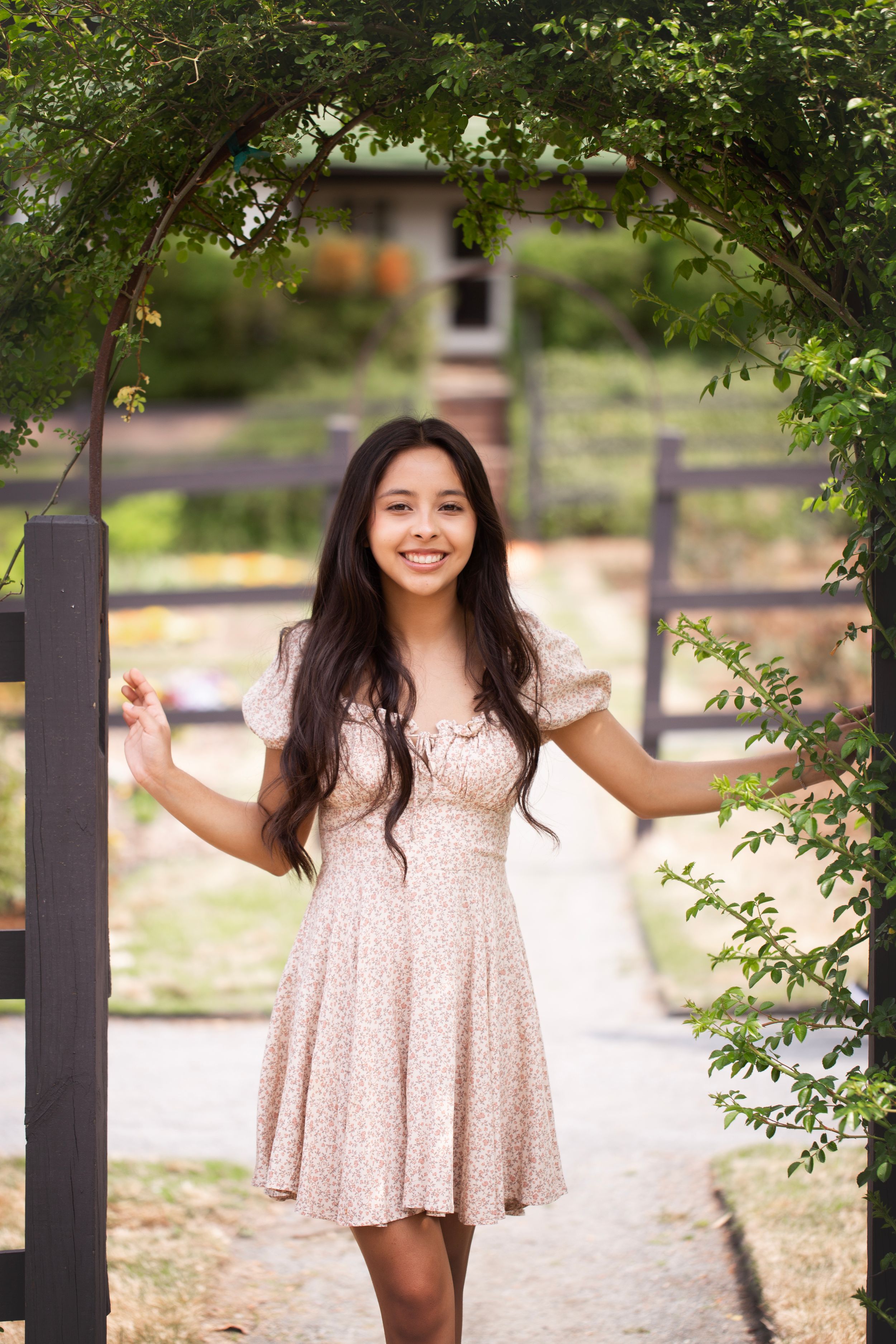 High school senior girl wearing a floral dress standing in a garden under a lattice archway.