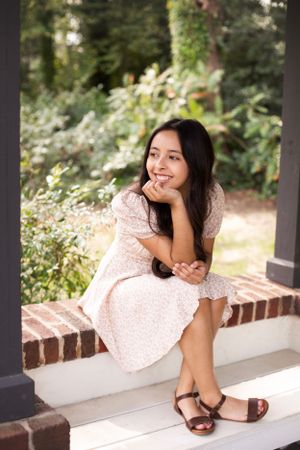 High school senior girl wearing a floral dress sitting in a gazebo in a garden looking off camera.