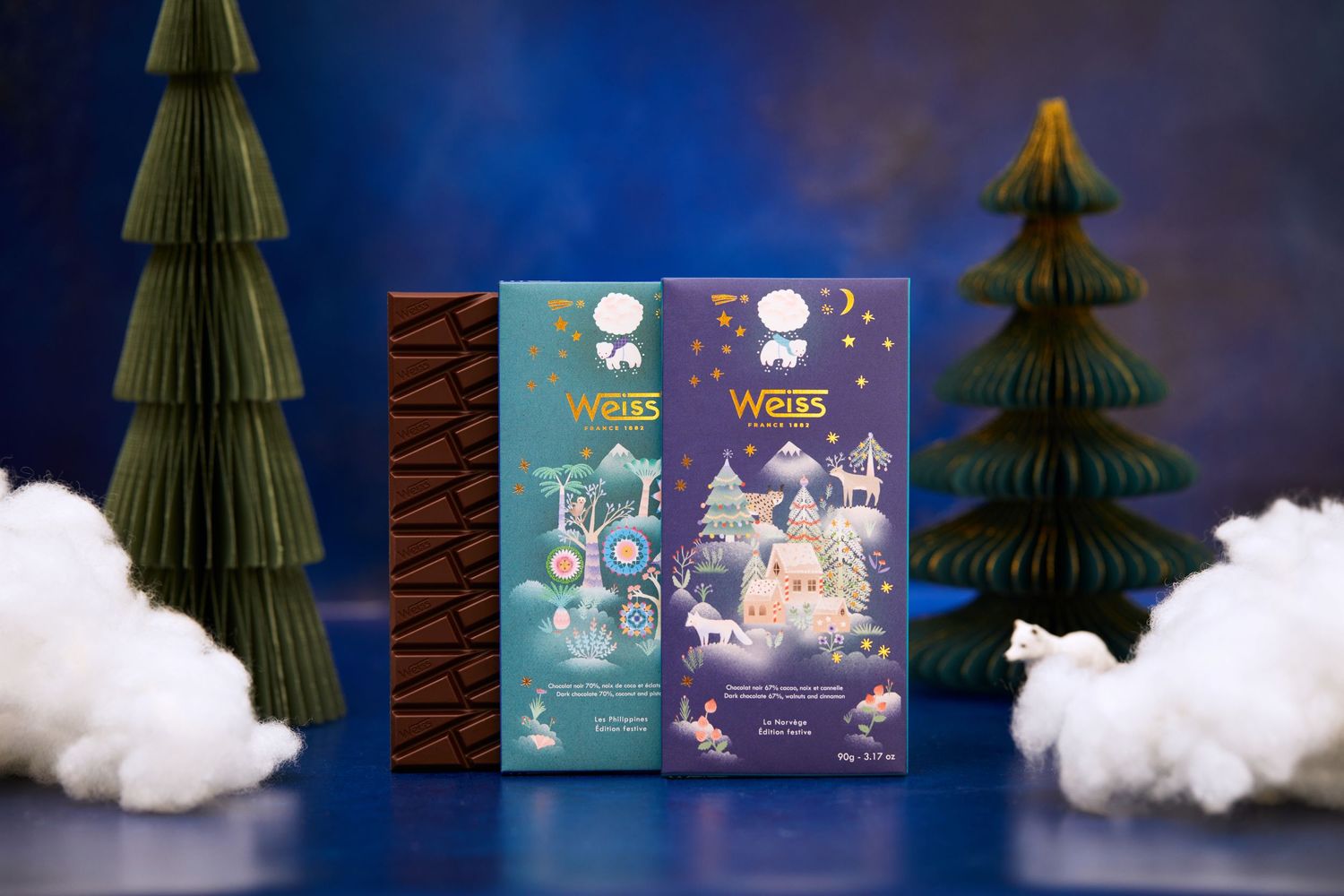 Two Weiss chocolate boxes displayed with decorative green paper trees and cotton cloud props against blue backdrop.