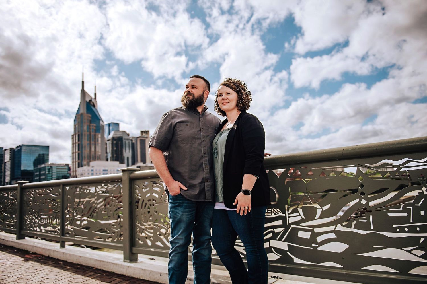 A couple stands on an urban bridge with skyline and clouds in background.