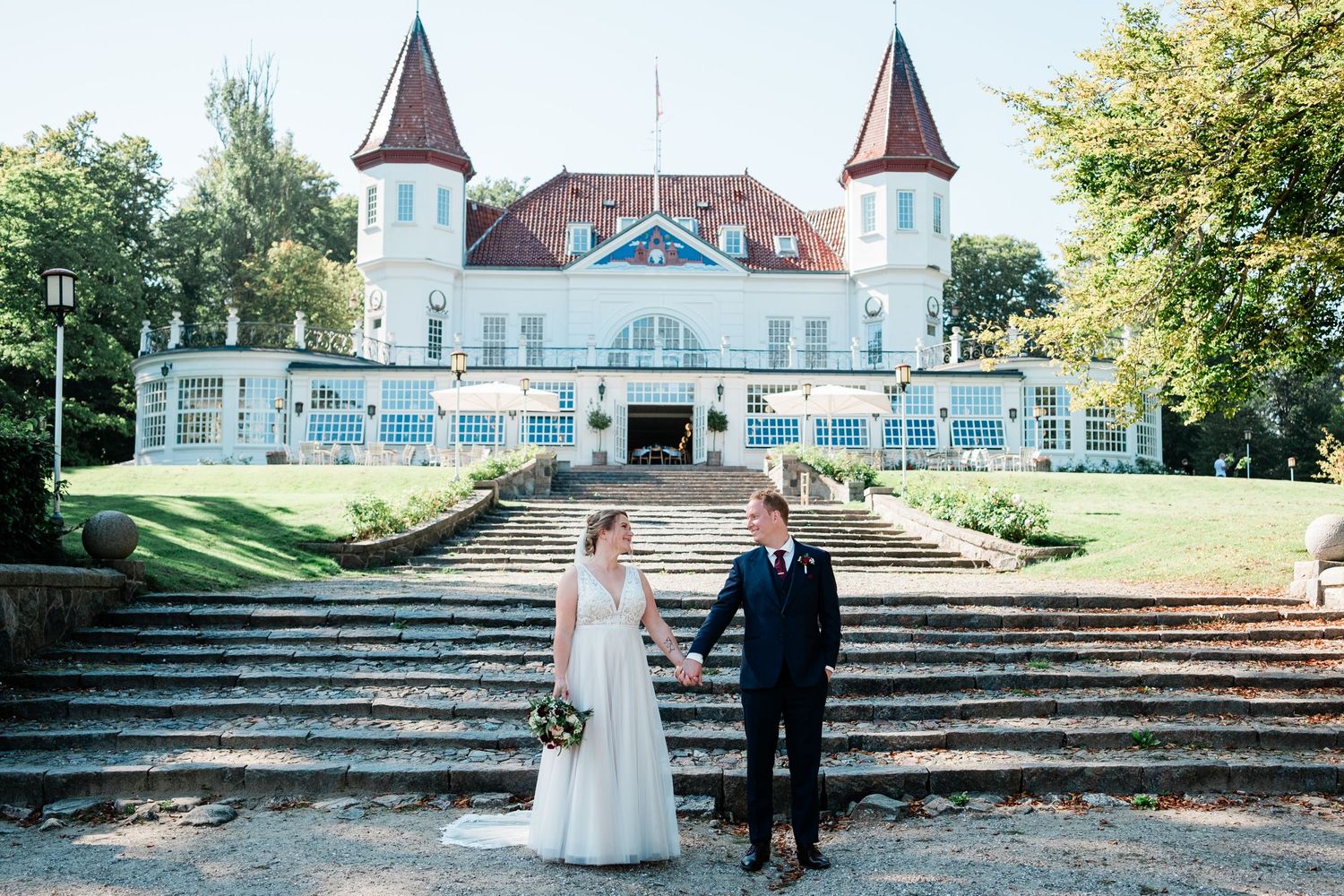 Elegant castle-like wedding venue with white exterior and twin towers behind couple on grand staircase.