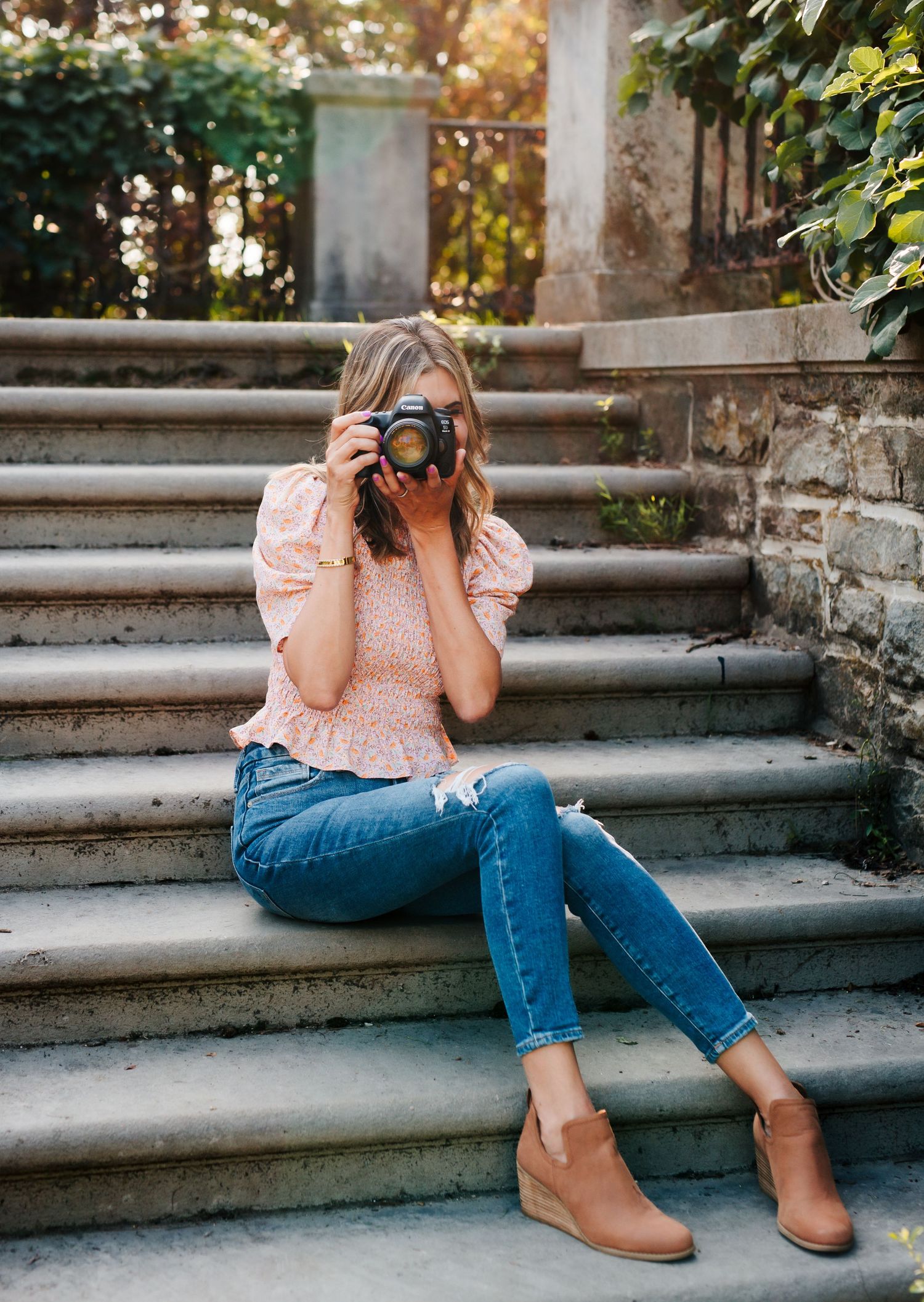 A photographer in ripped jeans and pink top sits on stone steps taking pictures.