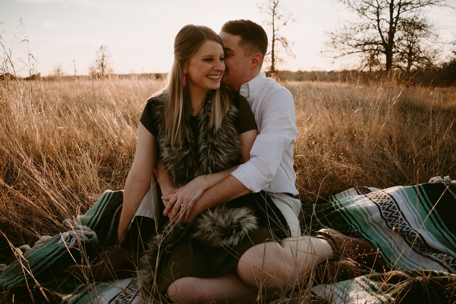 A couple sits intimately on a striped blanket in a golden field during sunset.