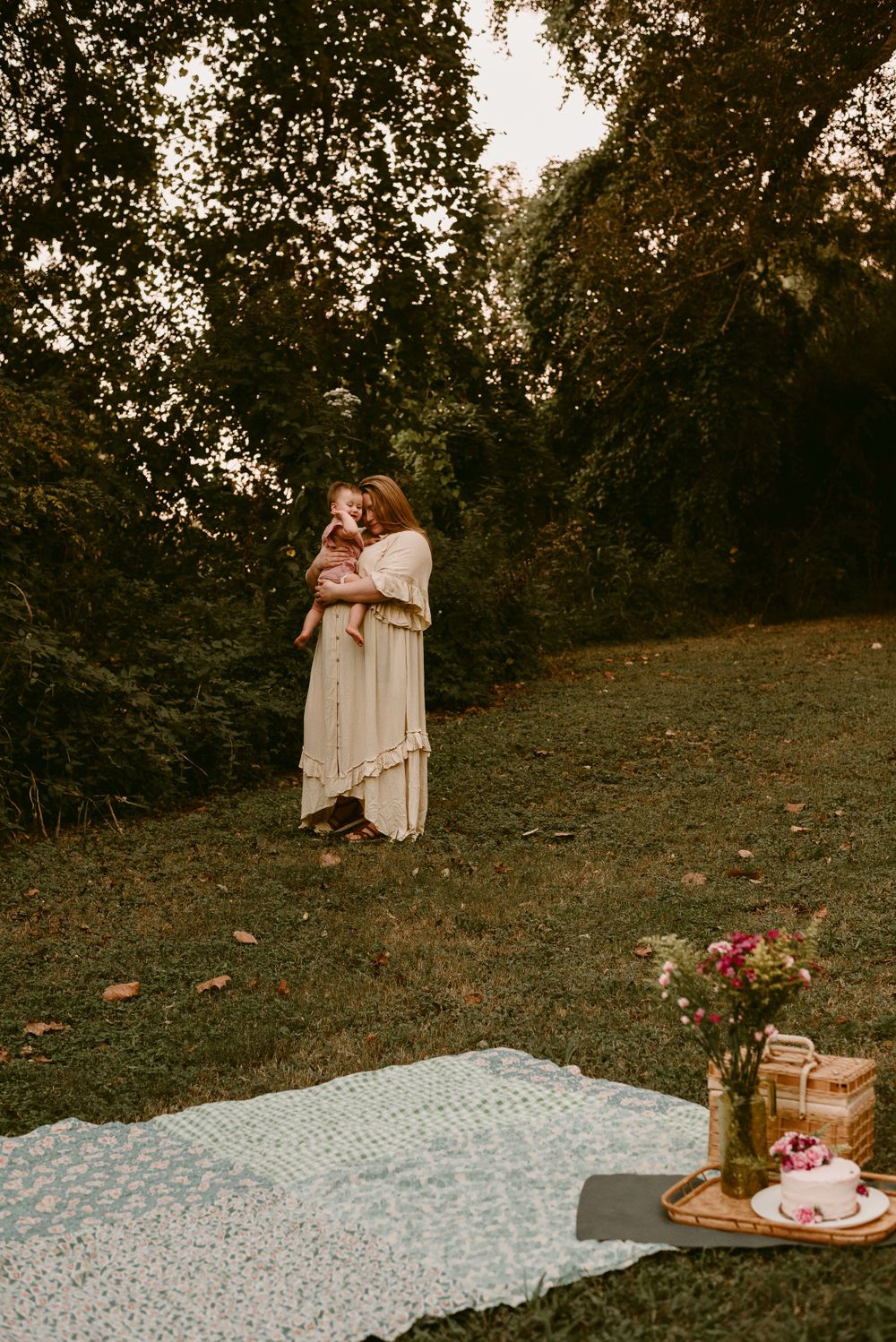 Dreamy outdoor scene with blanket picnic setup and figures standing among trees at dusk.