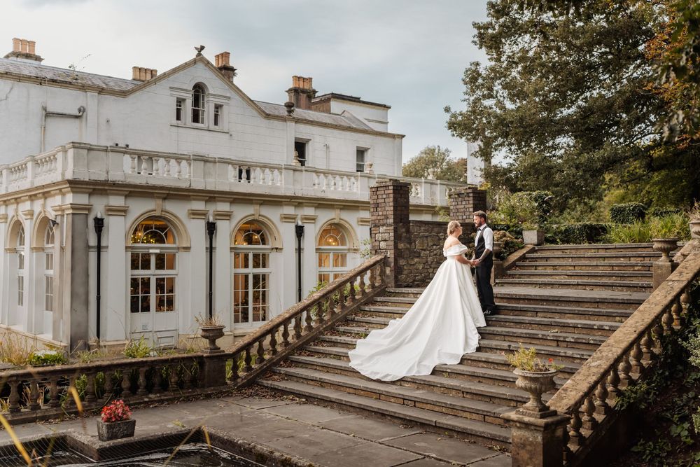 A romantic wedding couple poses on grand stone steps outside an elegant white mansion with ornate architecture.