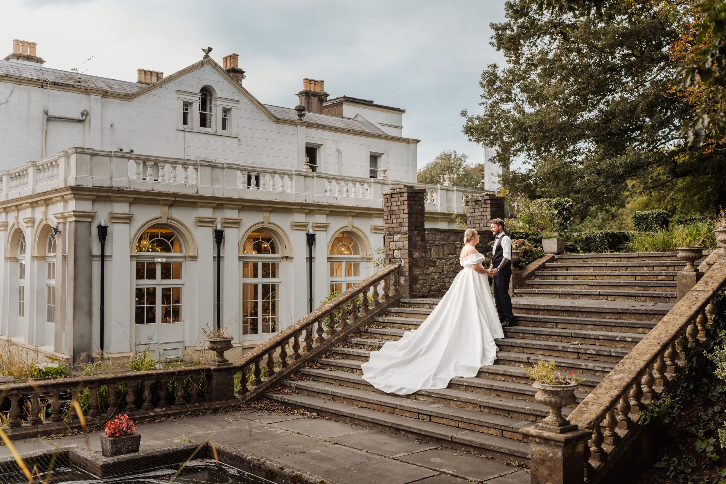A romantic wedding couple poses on grand stone steps outside an elegant white mansion with ornate architecture.