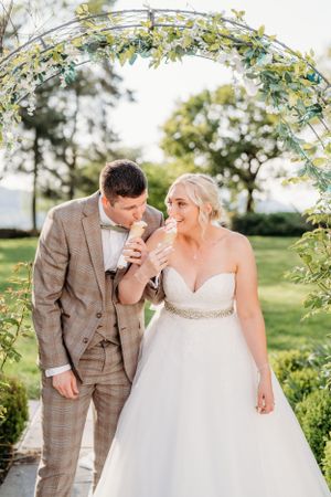 Couple shares ice cream under floral arch in garden wedding setting.