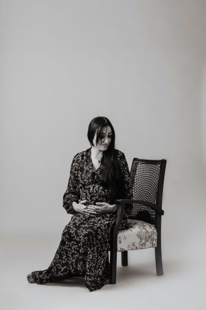 A moody black and white portrait of a woman in a floral dress seated beside an antique chair.