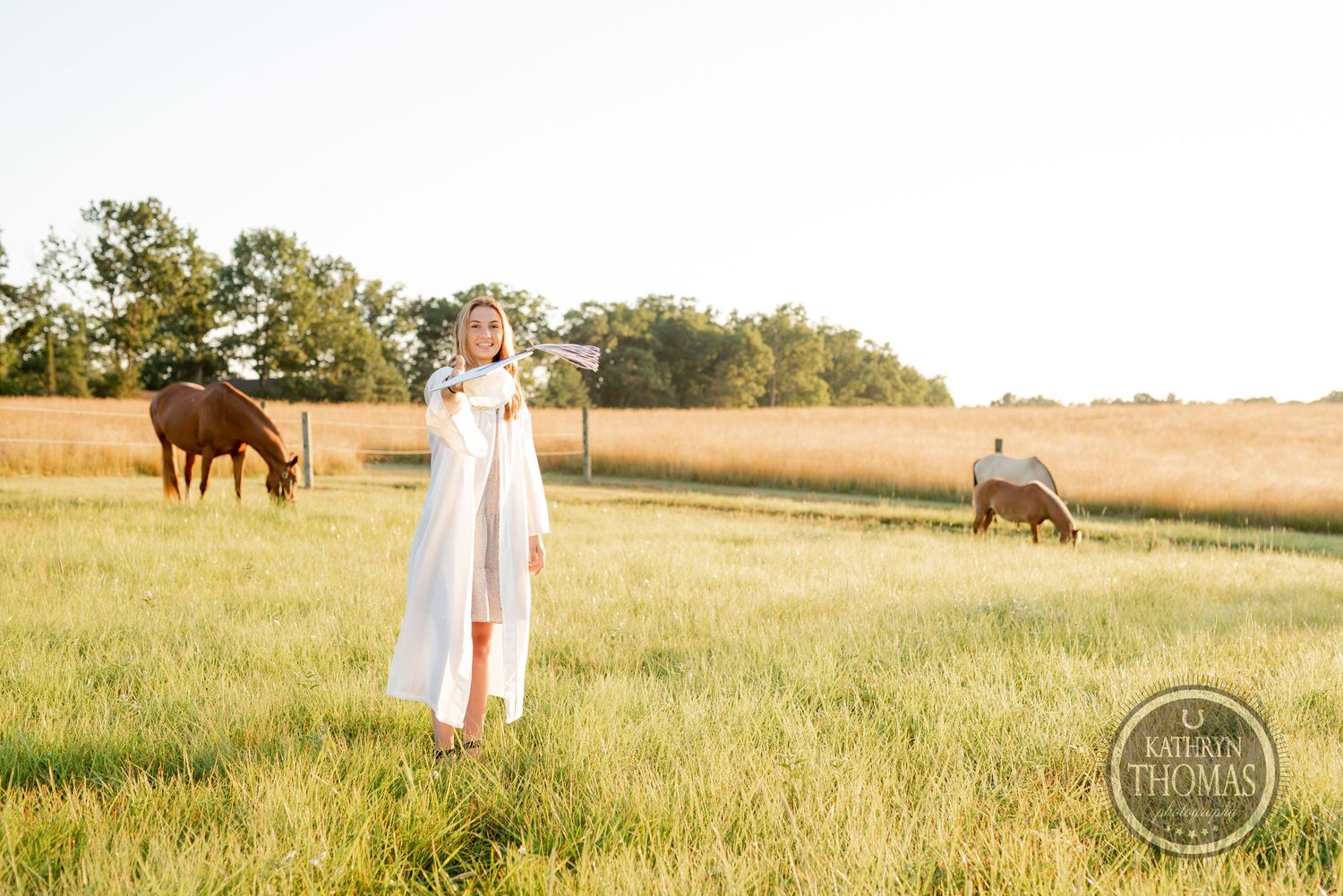 Graduate stands in flowing white gown in grassy field with horses roaming in background.