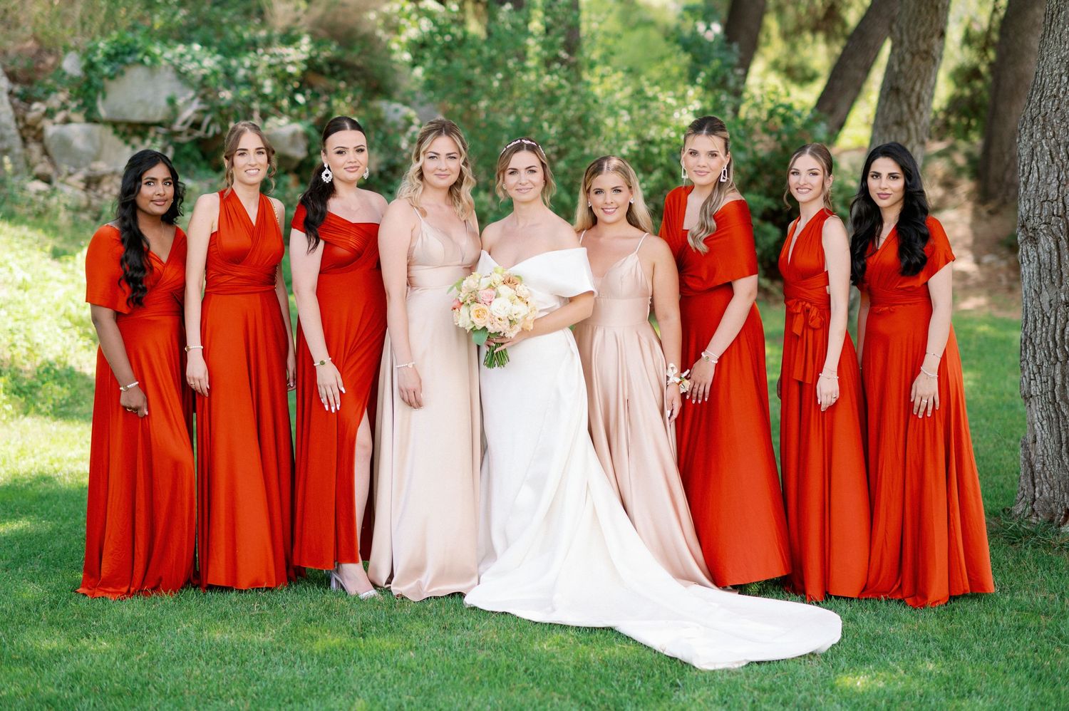 Wedding party wearing contrasting red and pink floor-length gowns posed together on grass in outdoor garden setting.