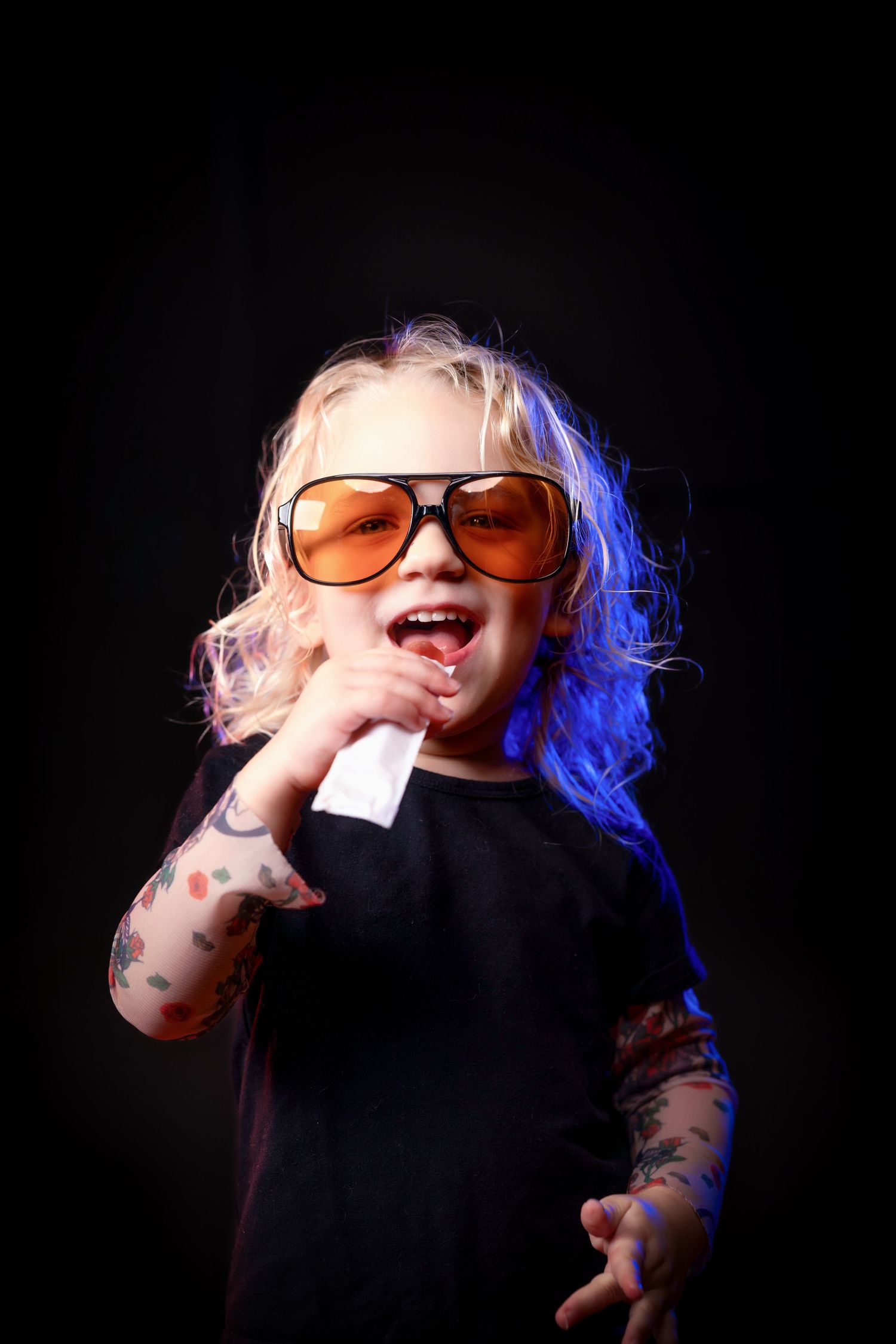 Adorable blonde toddler with fake tattoos wearing oversized orange sunglasses and black shirt under blue stage light.