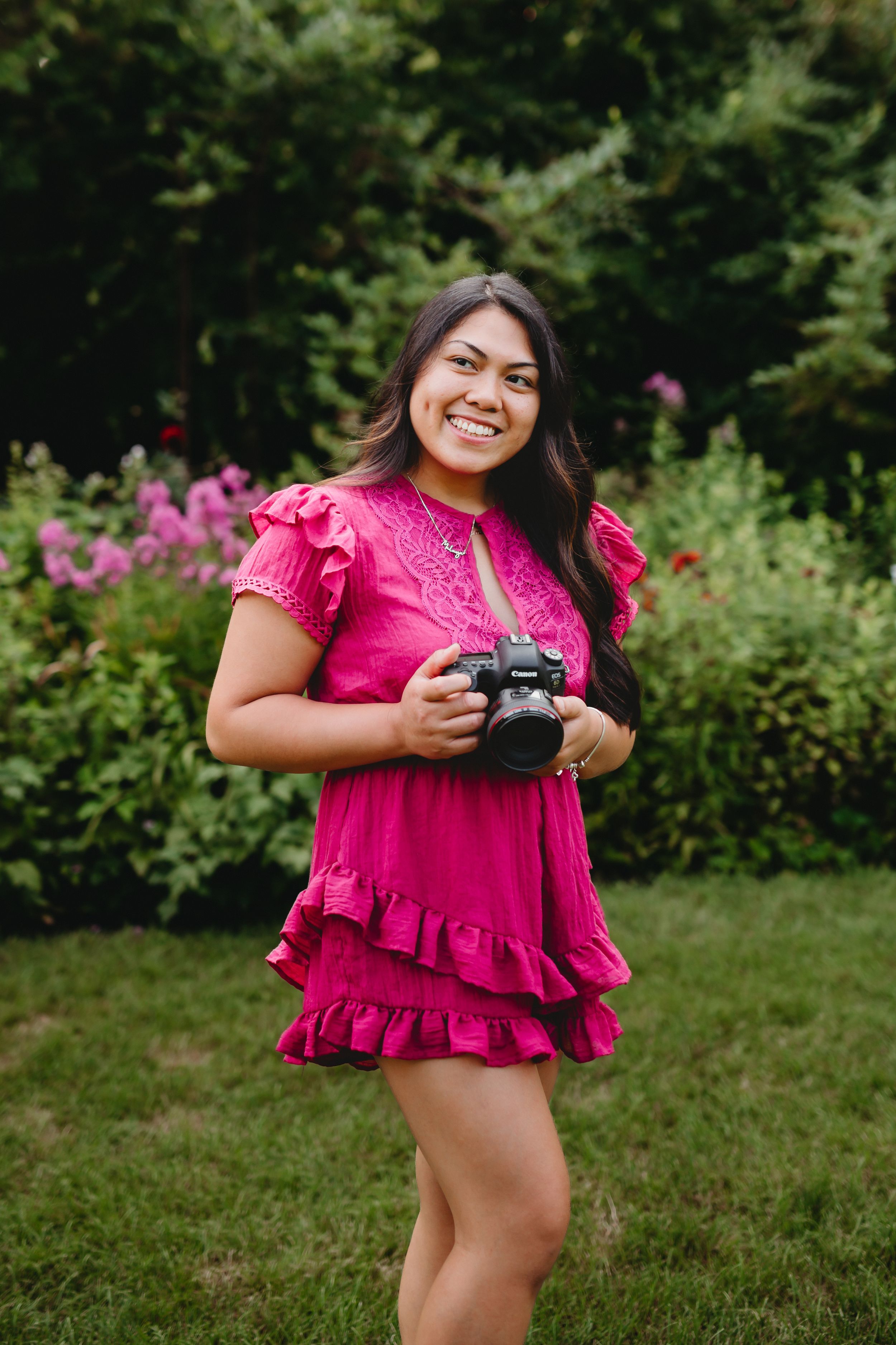 Woman in bright pink ruffle dress holds camera in blooming garden.