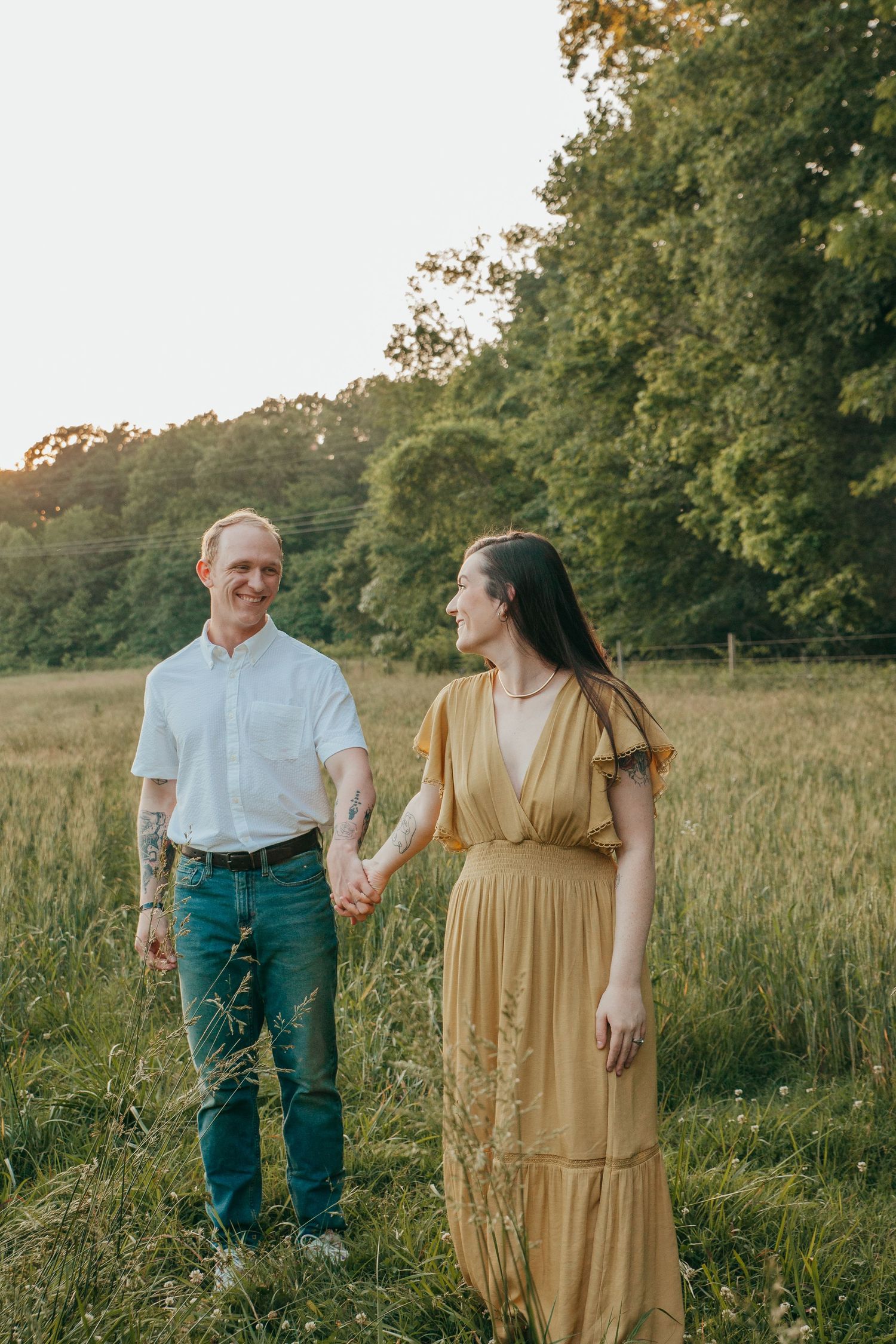 Couple in casual attire poses together in sunlit field at golden hour.
