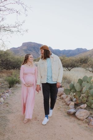 Two people walk hand in hand along a desert path lined with cacti and rocks.