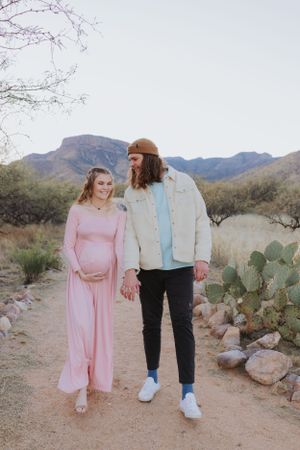 A couple strolls together on a desert trail with mountains and cacti creating a scenic backdrop.