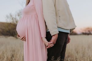 A couple holds hands in a field at sunset with a pink maternity dress and cream sweater.