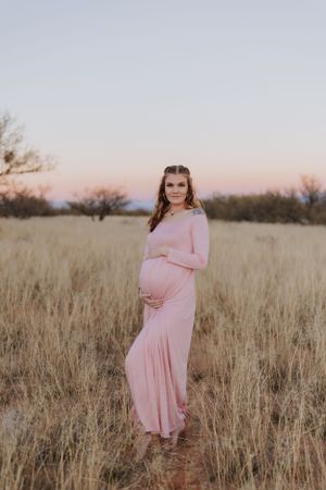 A maternity photo in a pink dress standing in a golden field at sunset.