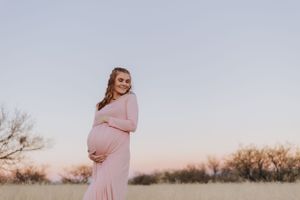 A maternity portrait in a flowing pink dress against dried grass and sunset sky.