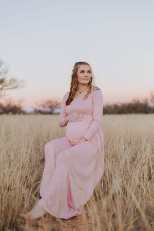 A maternity pose in a long pink dress surrounded by tall grass at dusk.