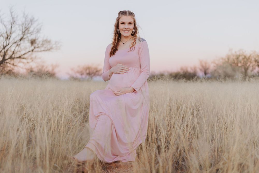 A maternity portrait in a pink maxi dress standing in golden grass.
