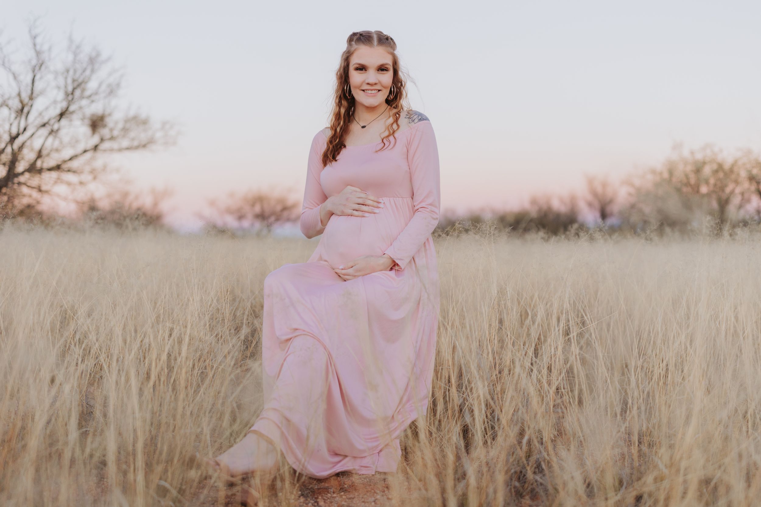 A maternity portrait in a pink maxi dress standing in golden grass.