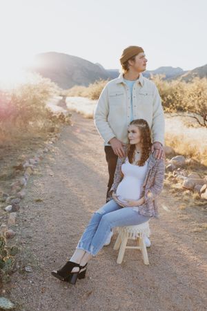 A couple sits together on a wooden chair along a desert trail at sunset.