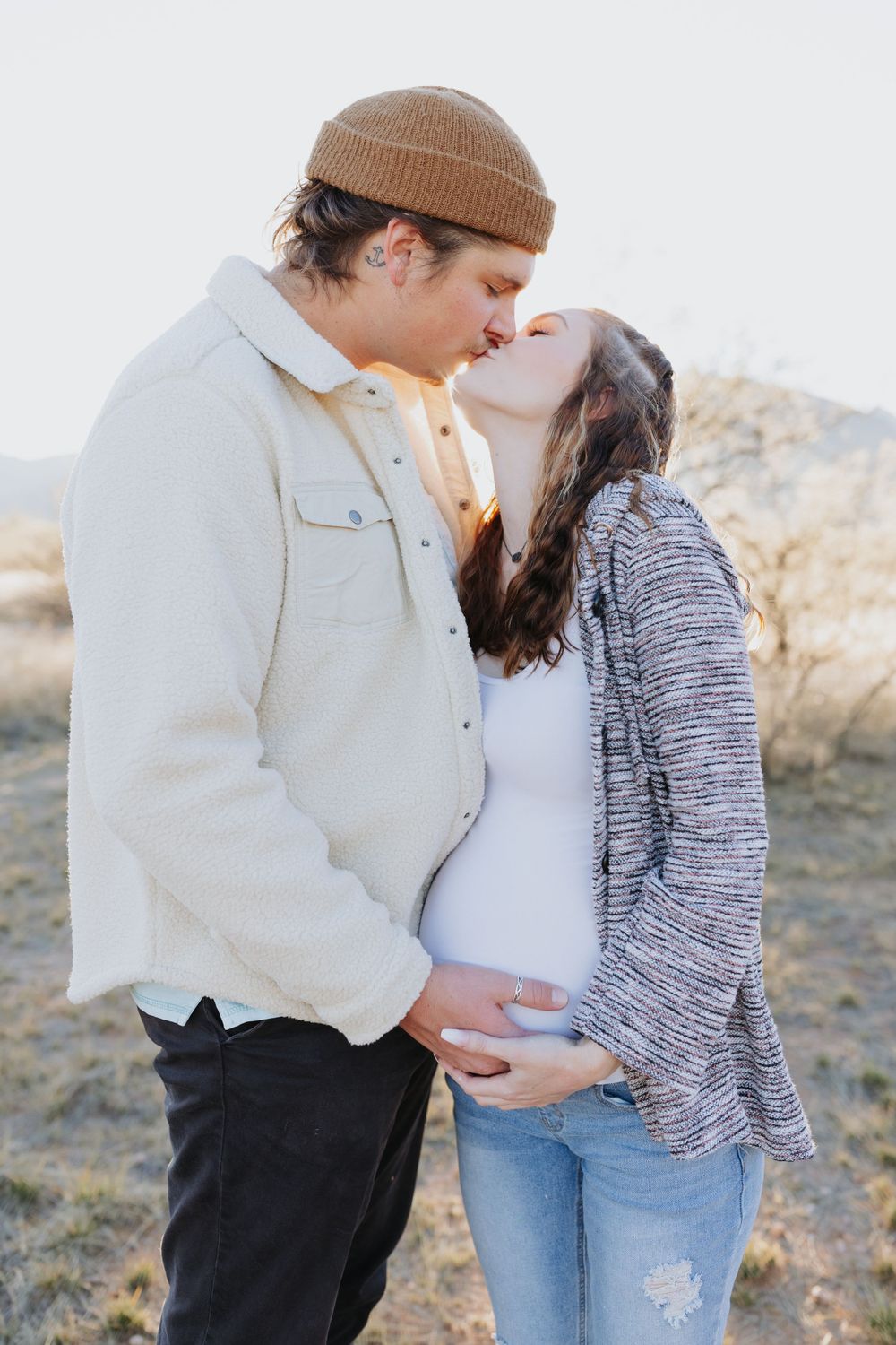 Couple in winter clothing embracing on desert hillside during sunset.