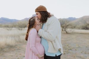 A couple embraces on a desert trail with mountains and brush in the distance.