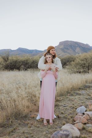 A couple in pink and cream outfits pose together in a desert setting with rocky mountains.