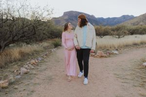 A couple poses together on a desert trail with mountains and cacti in the background during sunset.