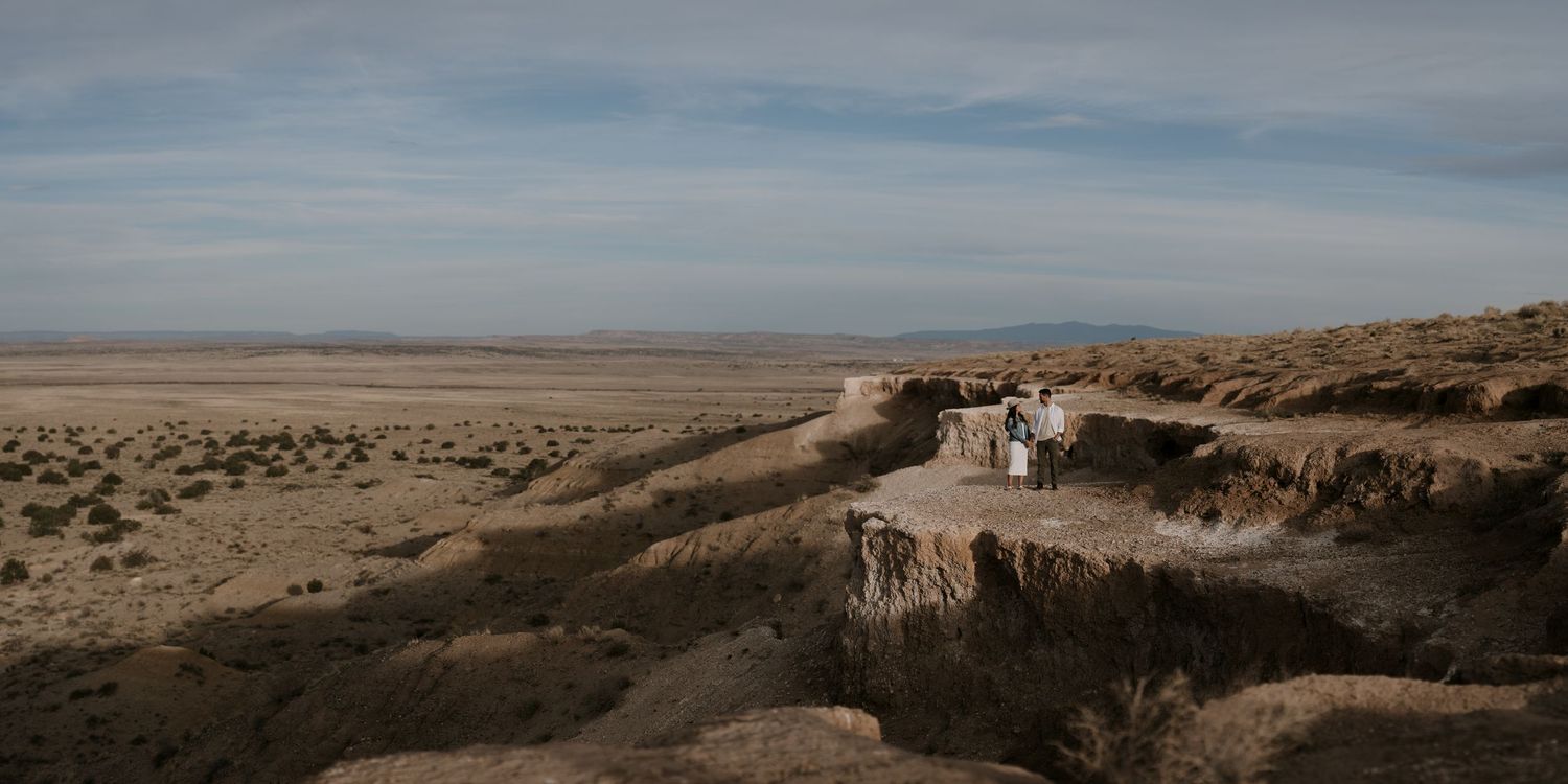 Distant aerial view of wedding couple standing on desert canyon edge under cloudy sky. by cinematic destination wedding photographers