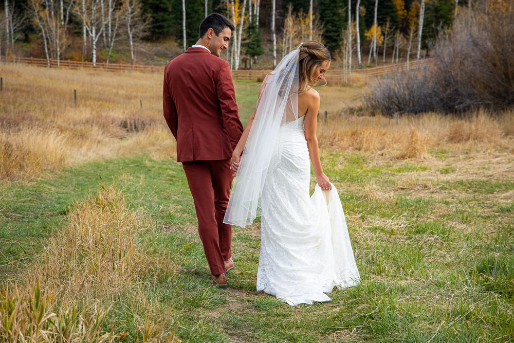 A couple in wedding attire walks through a field of tall grass with mountains in the background.