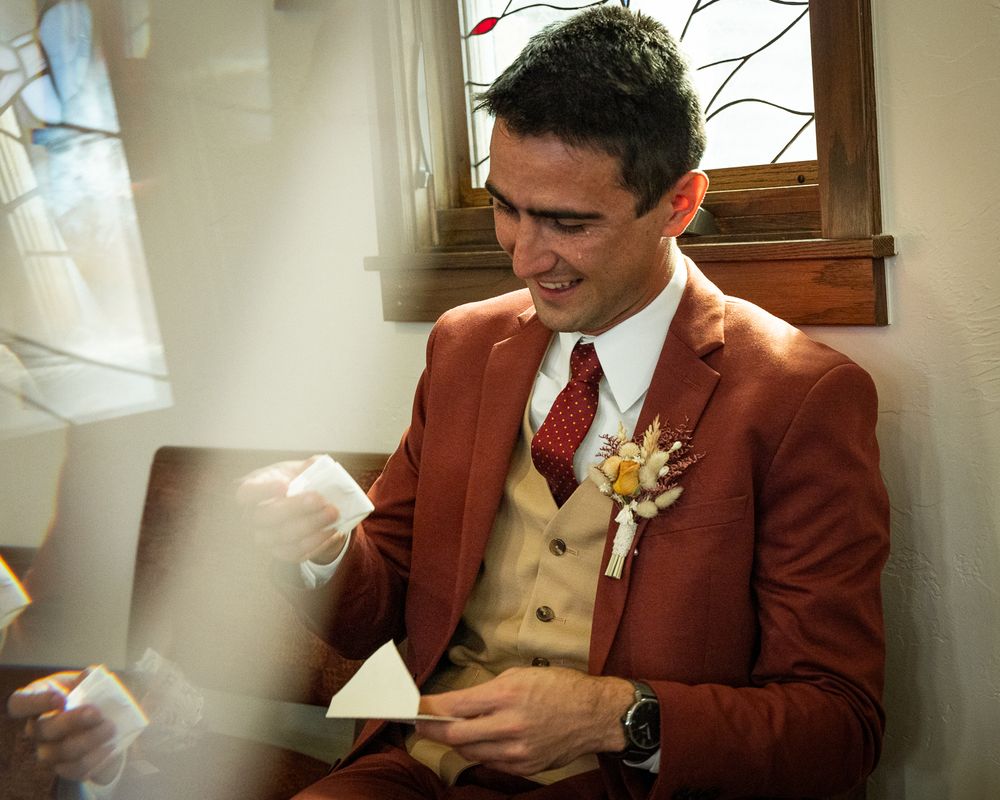 Groom in burgundy suit and tan vest reads letter while getting ready.