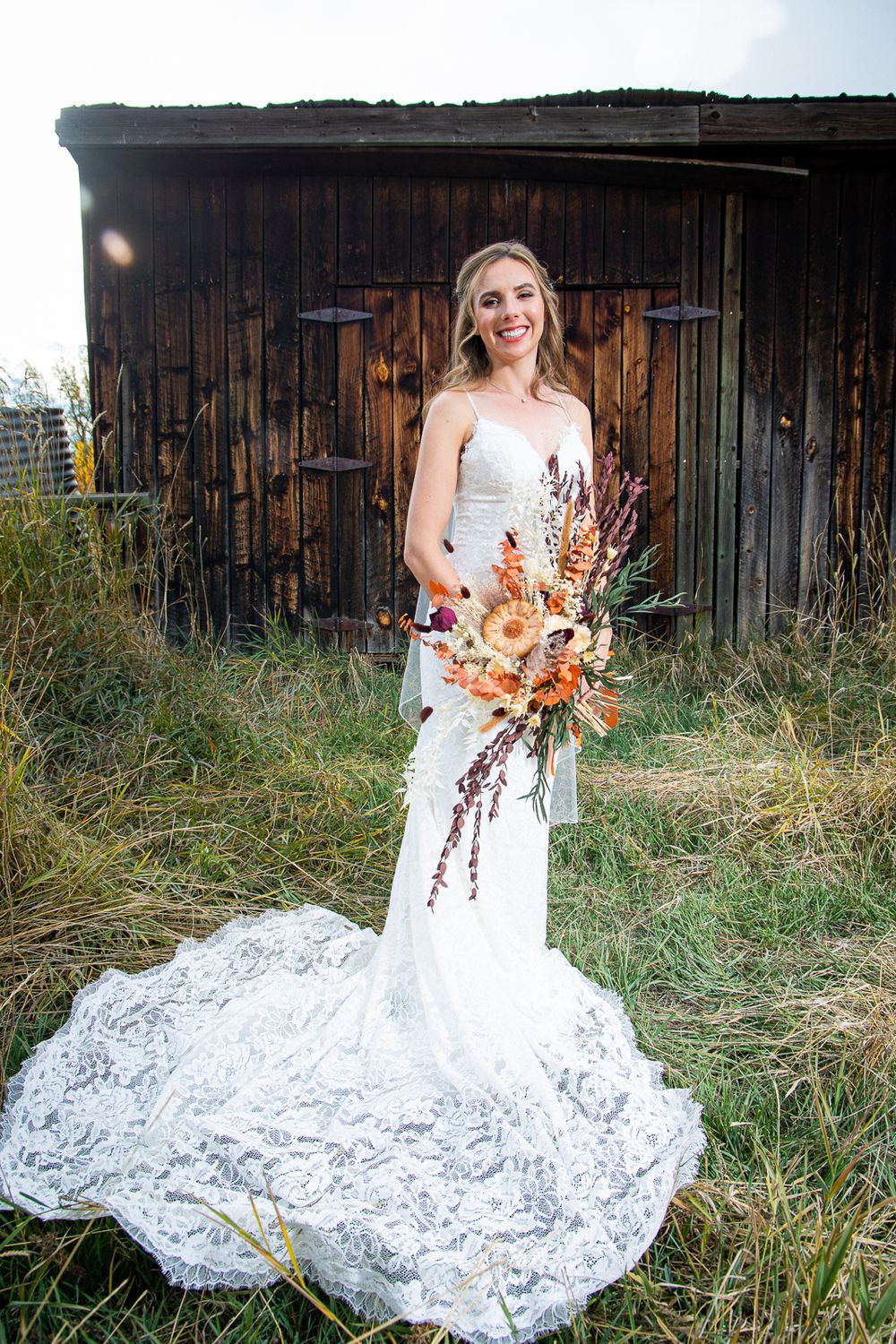 Bride in lace gown with trailing train holds orange and white bouquet against rustic barn.