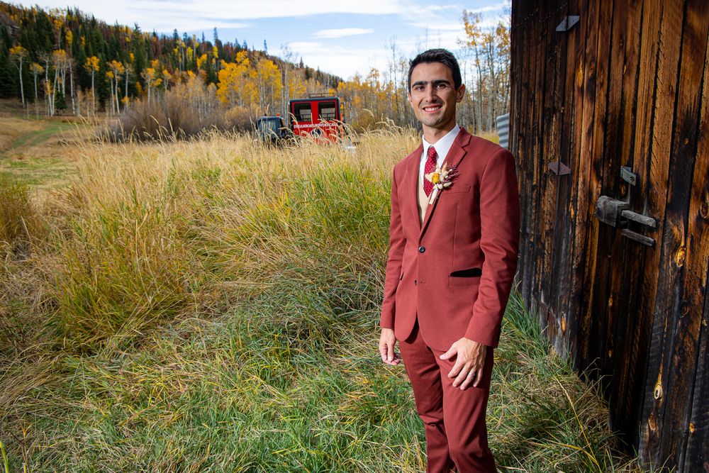 Groom in burgundy suit poses against rustic barn with autumn mountain landscape.