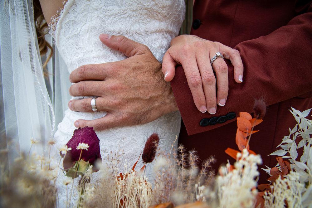 Close-up of hands showing wedding rings against a white lace dress and burgundy suit with flowers.