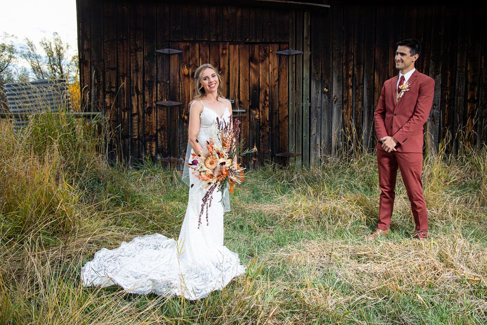 Wedding couple poses in front of wooden barn with autumn bouquet and burgundy suit.