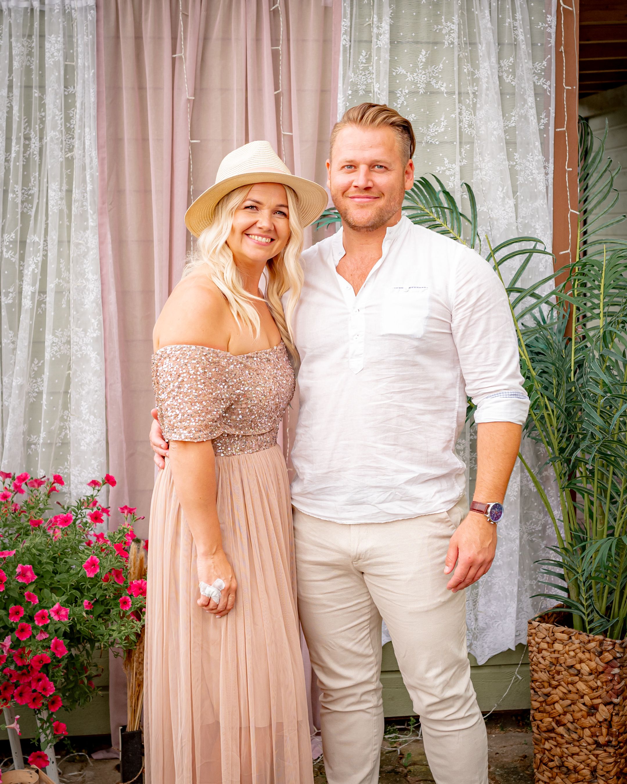 Couple in coordinated white and beige outfits pose together against a pink curtained backdrop.