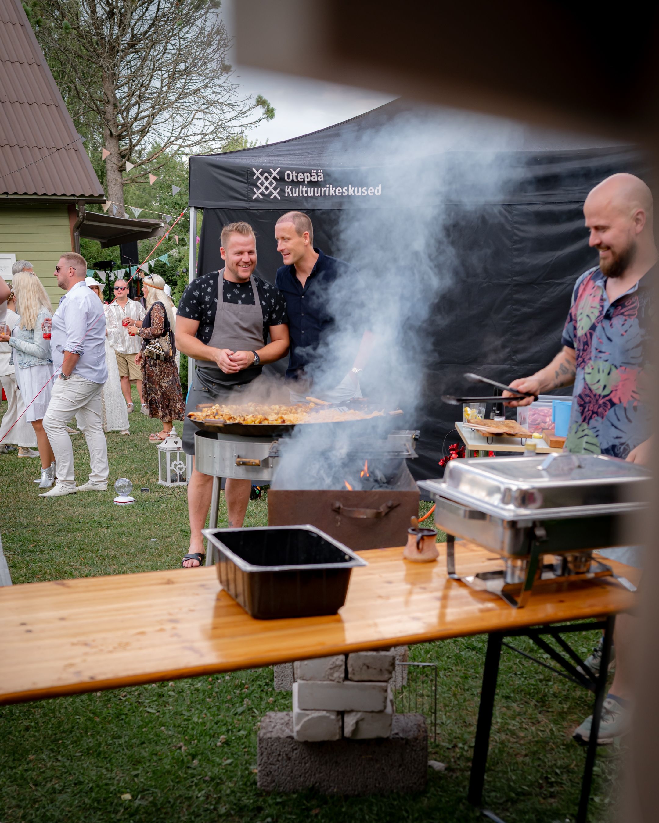 People grilling food at an outdoor event under a black tent canopy with smoke rising from the barbecue.