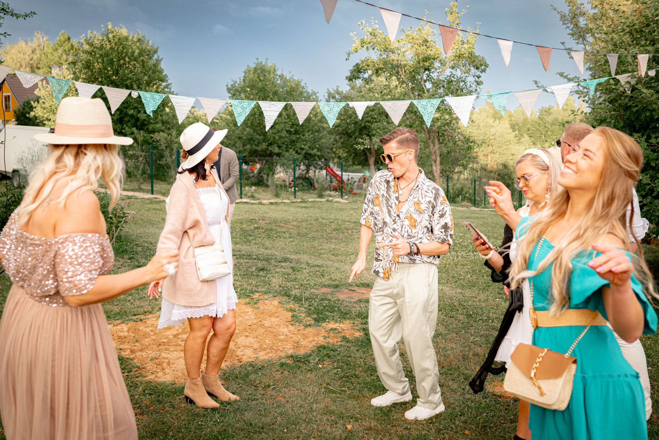Garden party guests in summer attire mingle beneath decorative bunting flags at an outdoor celebration.
