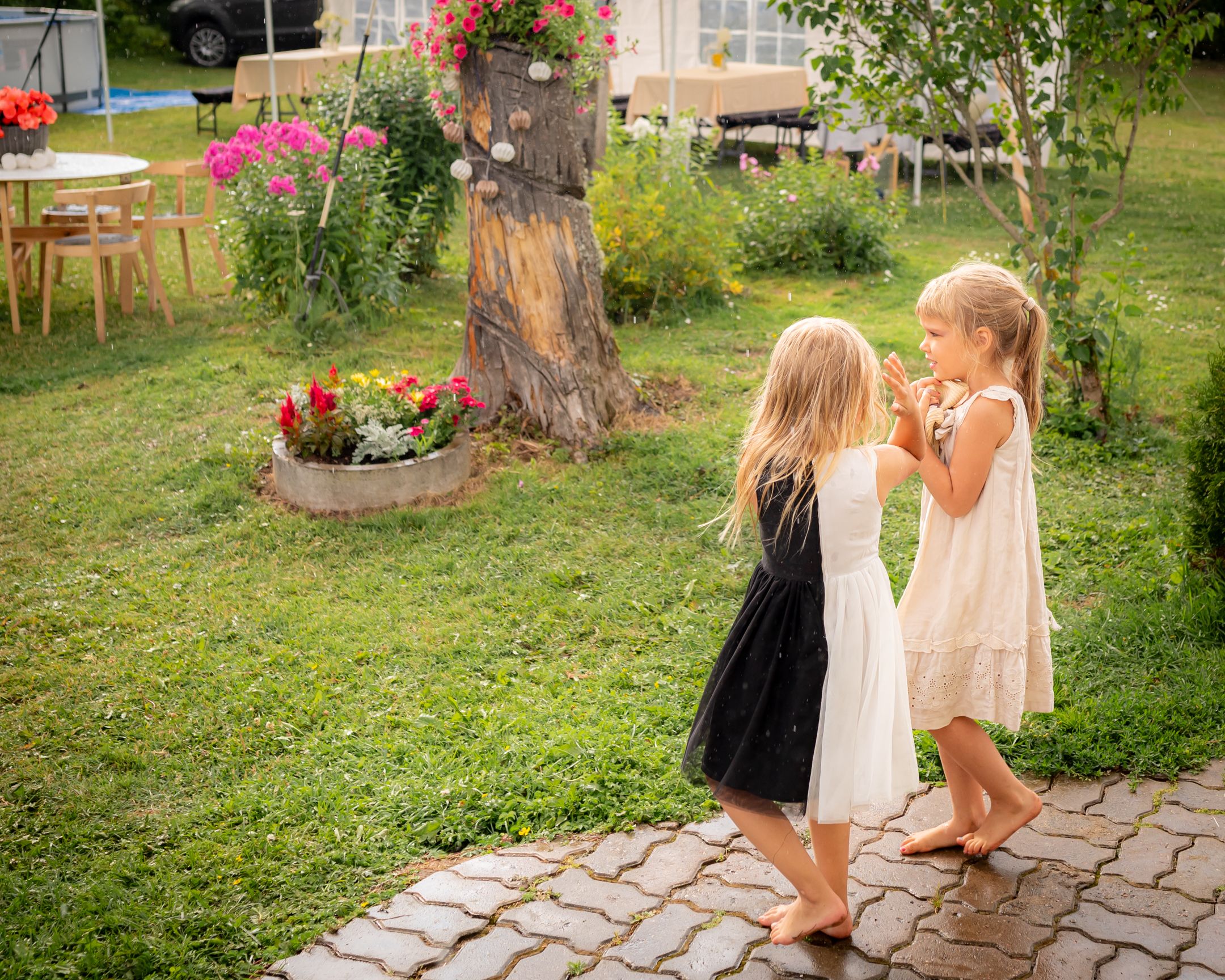 Two small children in dresses playing together on a stone patio with garden flowers in the background.