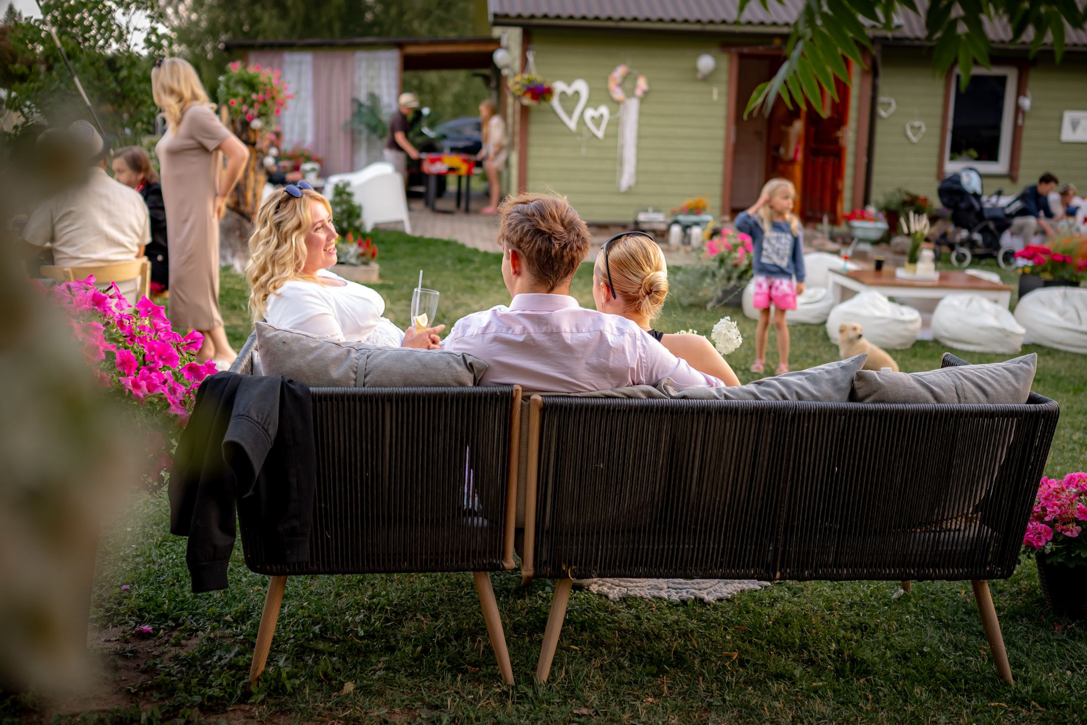 People relax on outdoor furniture during a backyard garden party with pink flowers.