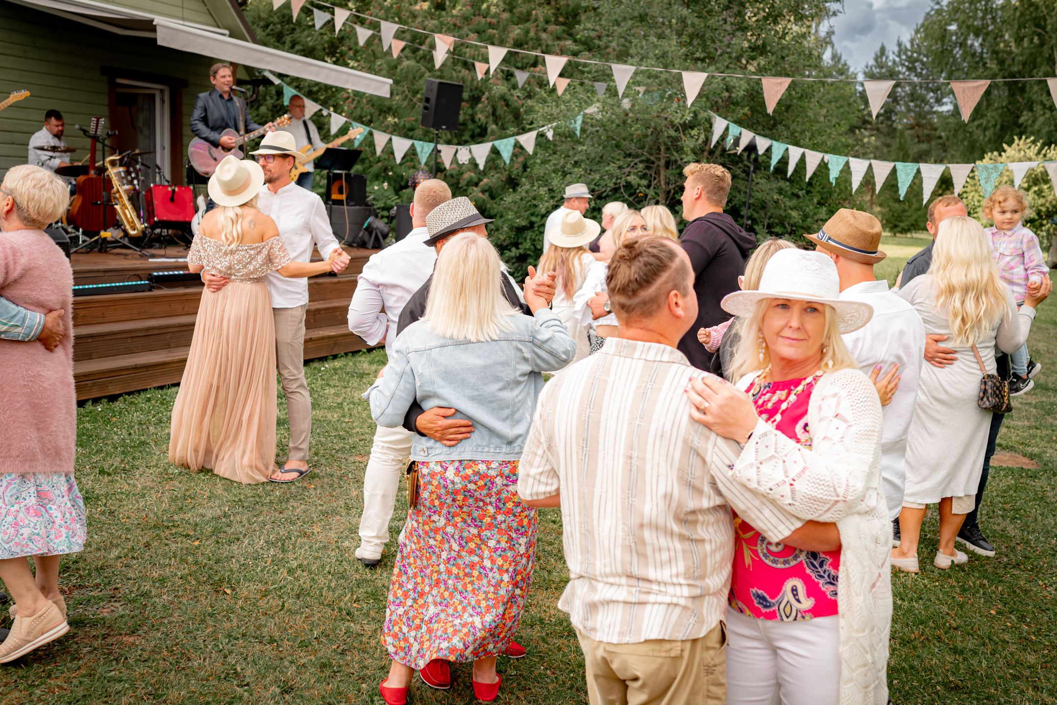 Couples dancing at an outdoor garden party with white bunting decorations overhead.