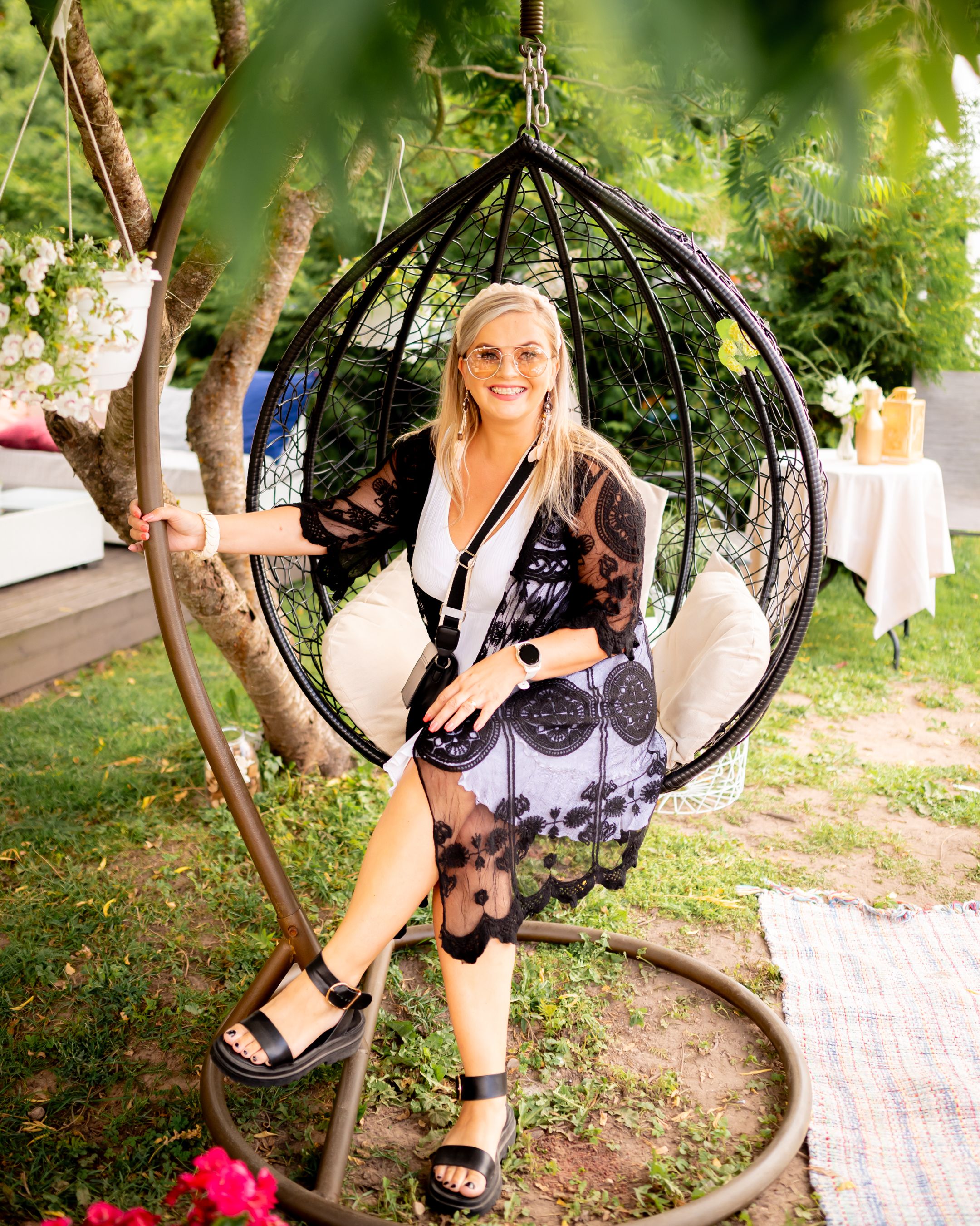 A person relaxes in a hanging egg chair surrounded by garden greenery and summer decor.