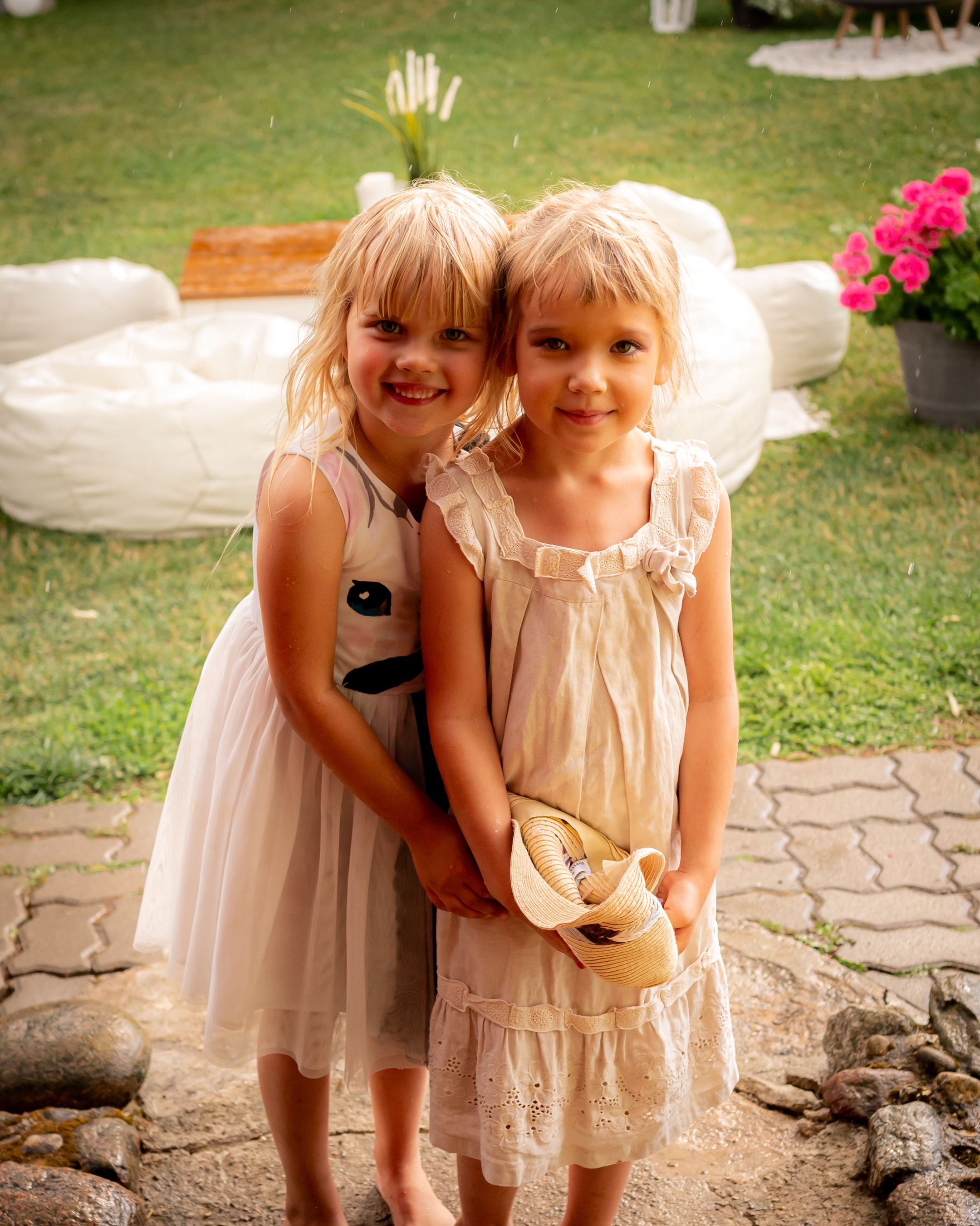 Two young children in light-colored dresses share a sweet moment together outdoors.