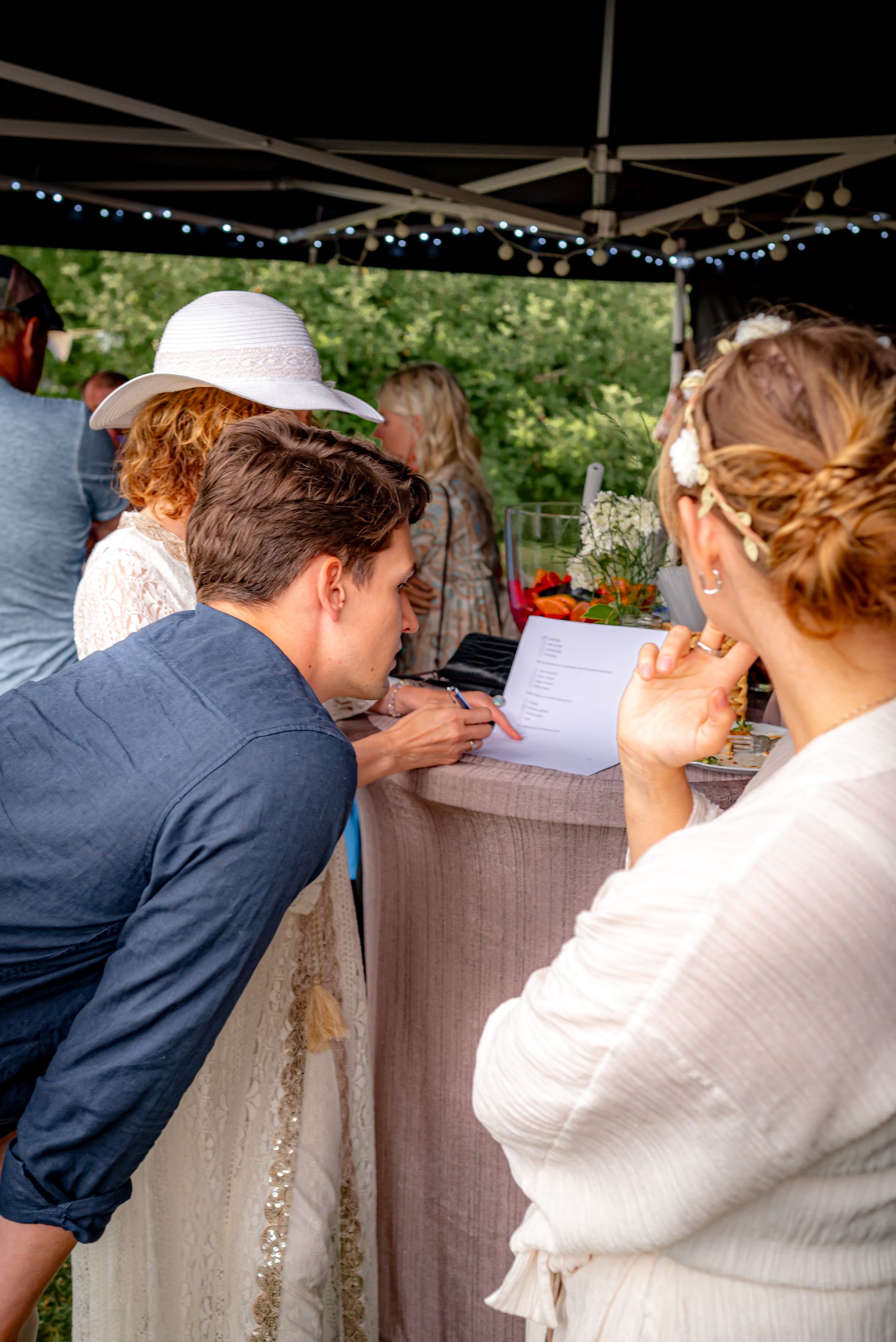 Attendees reviewing documents at an outdoor reception under a tent with string lights.